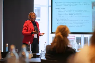 Cleopatra is in red blazer and speaks confidently at a conference. Behind her, a presentation slide discusses parent and community roles in schools. The audience is seated.