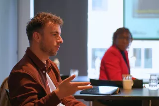 A man gestures while speaking in a meeting room; a woman listens attentively in the background. The setting is professional and focused.