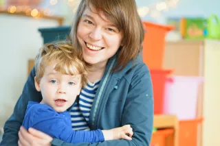 A smiling woman in a blue jacket embraces a young boy wearing a blue shirt. They are indoors, with colourful bins in the softly blurred background.