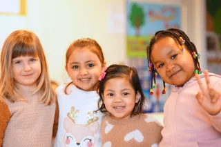 Four young children smiling in a classroom, wearing cozy sweaters. One child gestures a peace sign. The background has colourful artwork. Happy atmosphere.