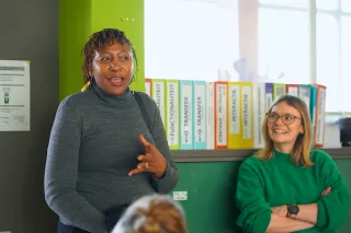 Woman in a gray sweater speaks animatedly in a bright room, while another woman in a green sweater smiles nearby. Colourful binders line the background, adding a lively tone.