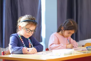 Two young girls focus on drawing at a classroom table. The girl on the left wears glasses and a colourful necklace, conveying a mood of concentration.