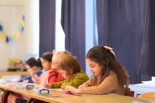 Children sit at a table in a classroom, focused on coloring activities. The room is softly lit, with dark curtains and a colourful garland in the background.