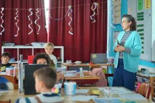 A teacher in a turquoise cardigan stands in a classroom with red curtains, talking to attentive students. The room is decorated with hanging spirals.