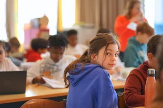 A young girl in a blue hoodie smiles at a classroom desk, surrounded by classmates with books and laptops. The atmosphere is studious and lively.