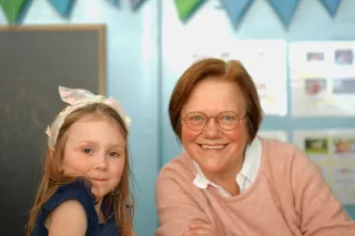 A young girl in a navy dress and headband smiles beside an older woman with glasses in a pink sweater. They sit in a colourful classroom, conveying warmth.