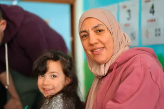 A woman in a pink hijab and a young girl smile warmly in a bright room. Educational posters with numbers are visible in the background.