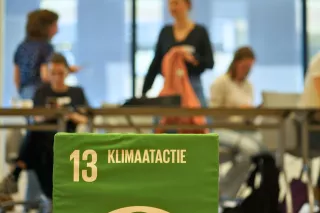 Green box with "13 Klimaatactie" in focus, symbolizing climate action. Blurry, diverse group of people in discussion in the background, indoors.