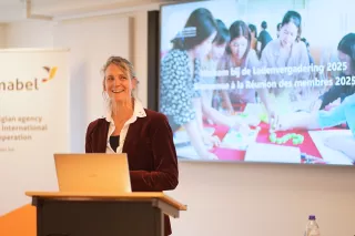 A woman stands smiling at a podium with a laptop. Behind her, a screen displays a group engaged in discussion, with text referencing a 2025 meeting.