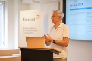 A man in a white shirt speaks at a podium with a laptop, next to a screen displaying "Review 2024." A banner for "Enabel" is in the background.