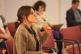 A woman in glasses speaks passionately while gesturing, seated among others blurred in the background. The scene conveys a focused discussion in a seminar.