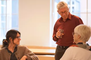 Three individuals in a bright room are having a conversation. A smiling woman in glasses sits facing a standing man holding a drink, both listening to another person.