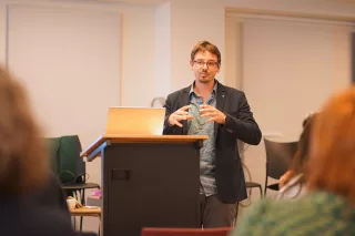 A man with glasses stands at a podium, speaking and gesturing with his hands. He appears engaged, with a laptop open in front of him. Chairs and blurred audience are visible in the background, suggesting an indoor presentation or lecture setting.