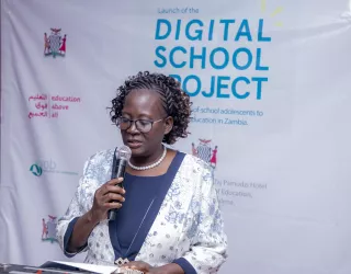 Woman speaking through a microphone, standing in front of a banner that says Digital School Project