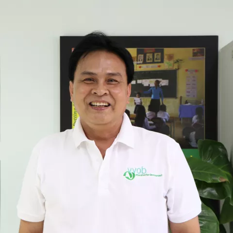 Vichetr wearing a white VVOB polo shirt, standing in front of a framed picture of students in a classroom.