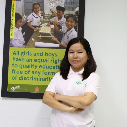 Srey wearing a white VVOB polo shirt, standing in front of a VVOB poster that says 'All girls and boys have an equal right to quality education, free of any form of discrimination.'