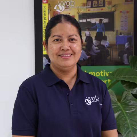 Chandaphea wearing a navy VVOB polo shirt, standing in front of a framed VVO picture of a classroom.