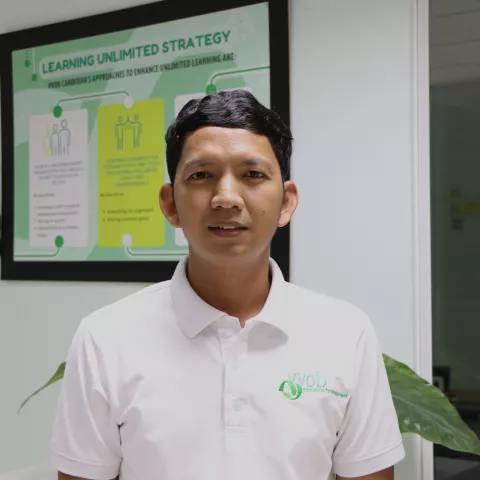 Phealay wearing a white VVOB polo shirt, standing in front of a frame VVOB banner that reads 'Learning Unlimited Strategy.'