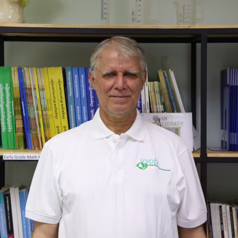Carl wearing a white t-shirt with the VVOB logo on it in green. He is standing in front of a bookshelf filled with blue and yellow books.
