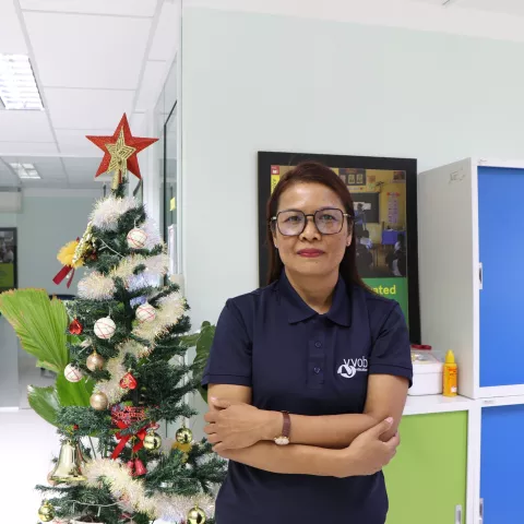 Tola wearing a navy VVOB polo shirt, standing next to a Christmas tree and colorful cabinets.