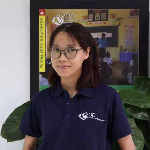 Sokkanha wearing a navy VVOB polo shirt, standing in front of a framed VVOB poster.