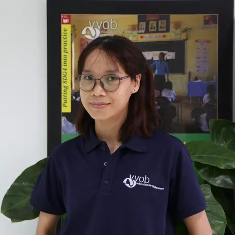 Sokkanha wearing a navy VVOB polo shirt, standing in front of a framed VVOB poster.