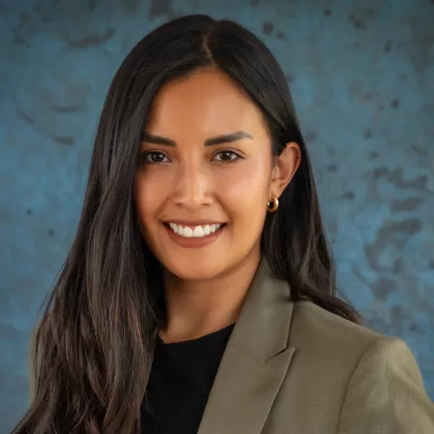 Alejandra smiles softly looking into the camera. She wears a light green blazer over a black shirt. The background is a mottled blue.