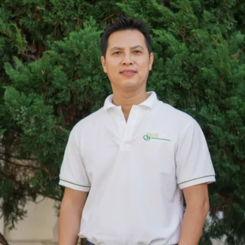 Chau  in a white polo shirt stands in front of lush green foliage, smiling softly. 