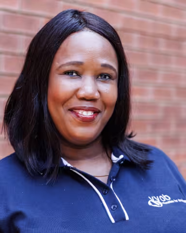 Ayande smiling in a navy polo shirt stands against a brick wall. Her expression is warm and friendly.