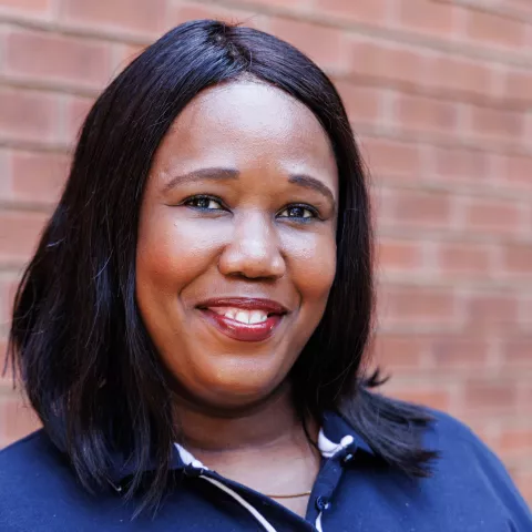 Ayande smiling in a navy polo shirt stands against a brick wall. Her expression is warm and friendly.