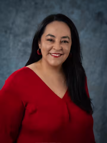 Carla smiles as the camera. She wears a red shirt. The background is a malted blue backdrop. 