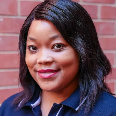Catherinah smiling in a navy polo shirt stands against a brick wall. Her expression is warm and friendly.