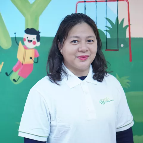Huyen white polo shirt smiles warmly in front of a colorful wall mural depicting a child reading and a red swing set.