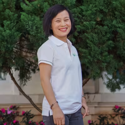 Ly smiles in a white VVOB polo shirt. She is standing in front of green plants and pink flowers.