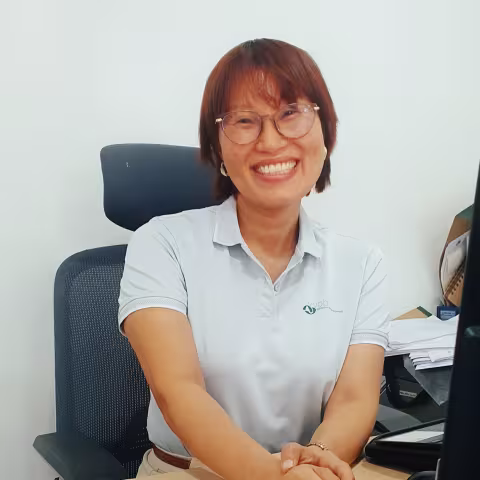Thin smiles warmly in a white VVOB polo shirt and glasses. She is sitting on a black office chair against a while wall. 