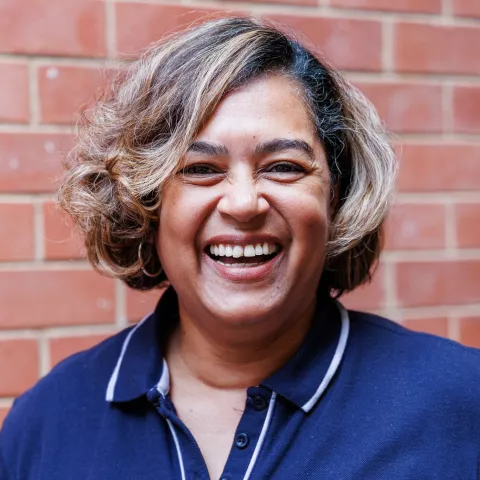 Chantal smiling in a navy polo shirt stands against a brick wall. Her expression is warm and friendly.