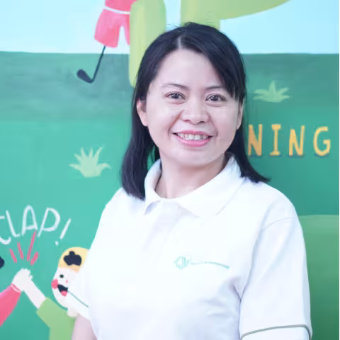 Tinh is wearing a white VVOB polo shirt. She smiles in front of a colorful mural featuring playful illustrations and words like "CLAP" and "NING." 