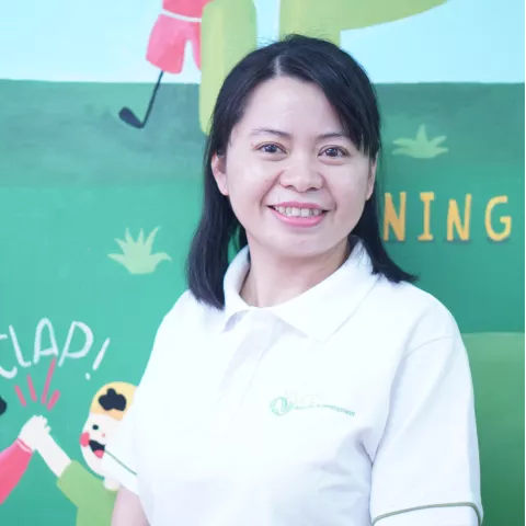 Tinh is wearing a white VVOB polo shirt. She smiles in front of a colorful mural featuring playful illustrations and words like "CLAP" and "NING." 