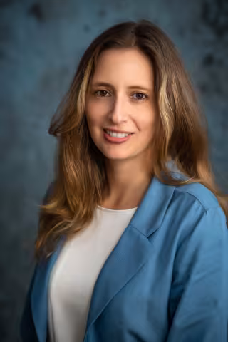 Claudia smiles softly looking into the camera. She wears a light blue blazer over a white shirt. The background is a mottled blue.