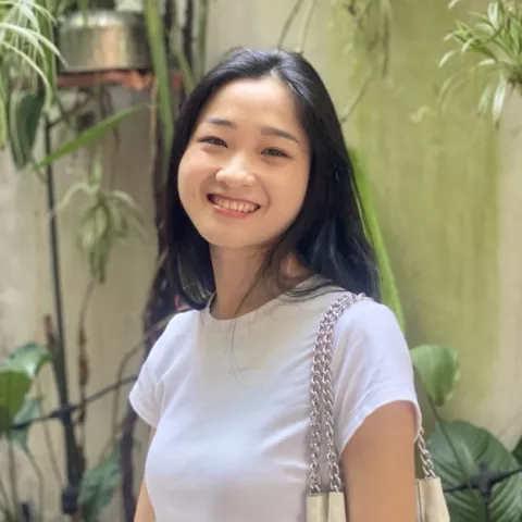 Diu is in a white shirt, holding a shoulder bag. She is standing in front of green plants, conveying a cheerful tone.