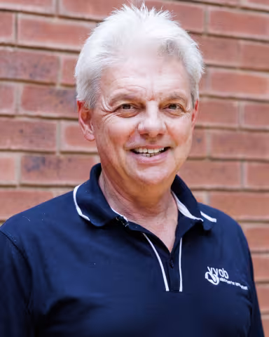 Wilfried smiling in a navy polo shirt stands against a brick wall. His expression is warm and friendly.