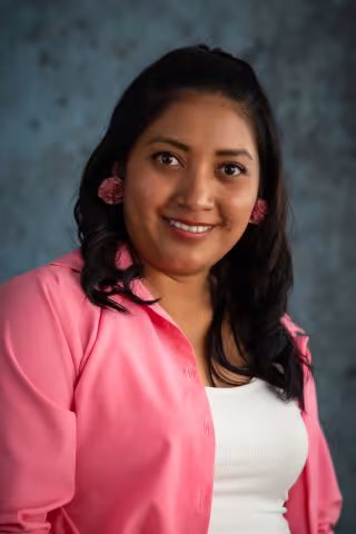 Evelyn smiles softly looking into the camera. She wears a pink blazer over a white shirt. The background is a mottled blue.