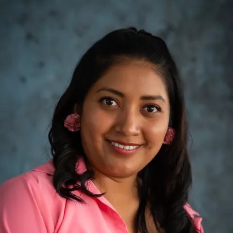 Evelyn smiles softly looking into the camera. She wears a pink blazer over a white shirt. The background is a mottled blue.