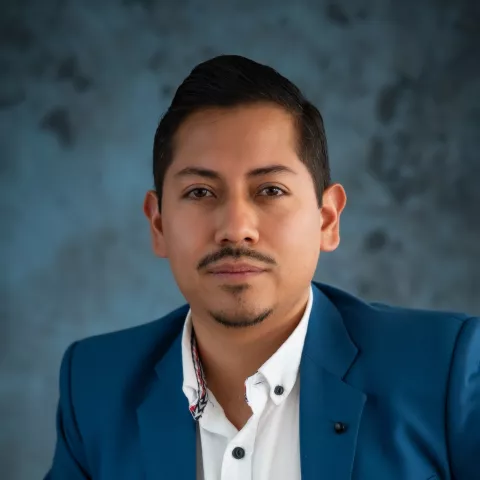 Fernando in a blue suit and white shirt poses confidently against a textured blue-gray backdrop. 