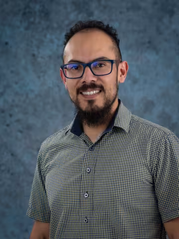 Francisco smiles at the camera. He wears glasses and a green and blue squared shirt. The background is a malted blue backdrop.