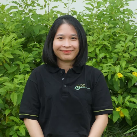 Hang is smiling, wearing a black VVOB polo shirt. She is standing in front of green plants.