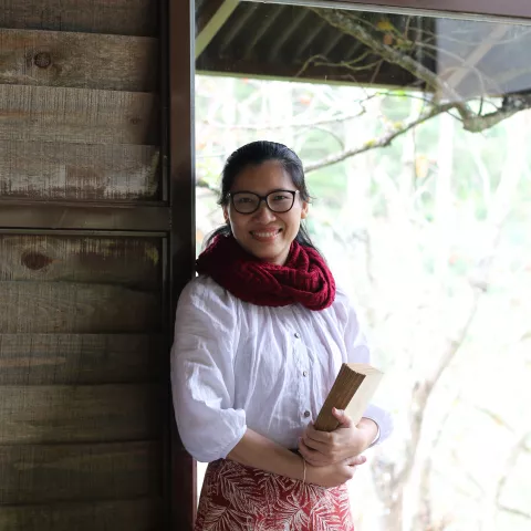 MaiAnh standing indoors by a window, holding a book. She wears glasses, a red scarf, a white blouse, and a patterned skirt. 