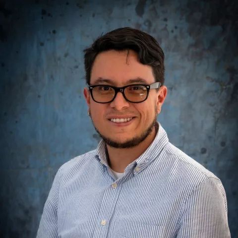 Jose smiles at the camera. He wears glasses and a blue shirt. The background is a malted blue backdrop.