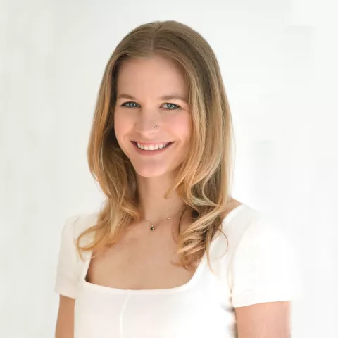 Kelsey smiles warmly at the camera. She is wearing a white shirt and standing in front of a white wall. 
