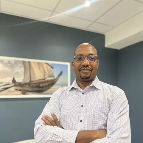  Kube is wearing a light blue shirt and glasses. He stands confidently with arms crossed in a living room. Behind him is a painting of a boat 